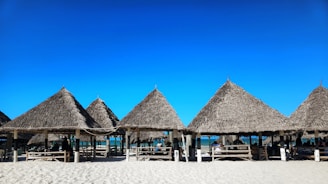 Several thatched-roof huts are arranged in a row on a sandy beach with a blue sky overhead. Wooden benches and tables are placed under the structures, and people are sitting in the shade, enjoying the seaside view.