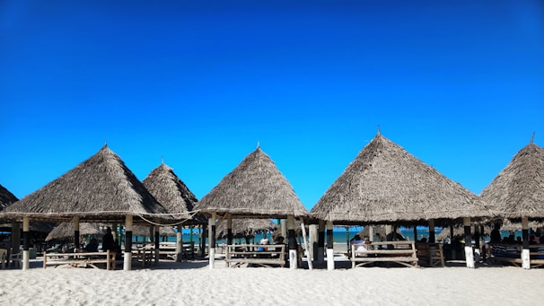 Several thatched-roof huts are arranged in a row on a sandy beach with a blue sky overhead. Wooden benches and tables are placed under the structures, and people are sitting in the shade, enjoying the seaside view.