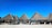 Several thatched-roof huts are arranged in a row on a sandy beach with a blue sky overhead. Wooden benches and tables are placed under the structures, and people are sitting in the shade, enjoying the seaside view.