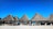 Several thatched-roof huts are arranged in a row on a sandy beach with a blue sky overhead. Wooden benches and tables are placed under the structures, and people are sitting in the shade, enjoying the seaside view.