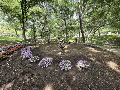 A scenic garden area is adorned with vibrant flowers, including purple and pink blossoms, set against a backdrop of lush, green trees. In the center, a sculpture of a playful child balances on one hand, capturing the serene and whimsical nature of the setting.