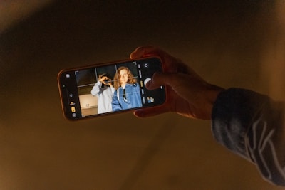 A person holding a smartphone with the camera app open, capturing a photo of two individuals standing close together. One person is making a peace sign, and the other is smiling. The image is taken in a dark environment.