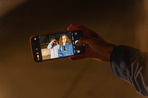 A person holding a smartphone with the camera app open, capturing a photo of two individuals standing close together. One person is making a peace sign, and the other is smiling. The image is taken in a dark environment.