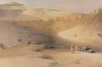 A vast desert landscape with rocky cliffs surrounding a flat valley. A small group of people are gathered on the ground, some sitting and others standing, dressed in traditional attire. The earth tones of the sand and rocks dominate the scene, with gentle gradations of light and shadow.