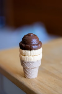 A toy ice cream cone with a brown and beige scooped top made of plastic, placed on a wooden table. The ice cream cone has a waffle pattern on the base and displays a layered appearance of chocolate and vanilla scoops.