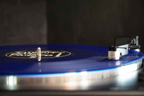 A close-up view of a blue vinyl record spinning on a turntable. The needle arm is positioned on the edge of the record, ready to play. The label on the record features white text on a black background. The scene is lit softly, highlighting the glossiness of the vinyl.