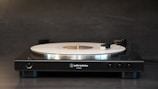 A sleek, modern turntable is positioned against a dark background, featuring a white vinyl record on its platter. The tonearm is slightly raised, and the turntable bears the branding of Audio-Technica.