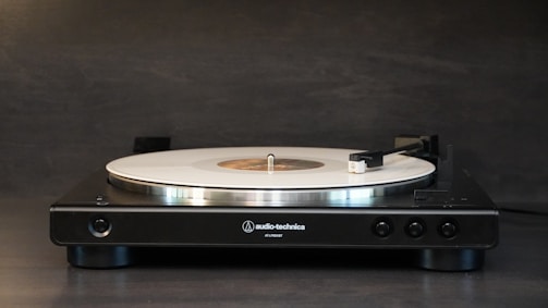 A sleek, modern turntable is positioned against a dark background, featuring a white vinyl record on its platter. The tonearm is slightly raised, and the turntable bears the branding of Audio-Technica.