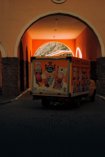 A delivery truck with colorful advertisements for ice cream is parked beneath a stone archway. The warm lighting inside the arch contrasts with the darker shadows outside. The truck features various ice cream visuals and text, creating a vibrant display. In the background, partially visible rooftops are illuminated by warm light, adding depth to the scene.
