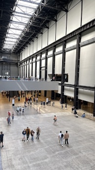 A high-ceilinged industrial indoor space with a large open floor area. The building structure features high walls with a series of tall vertical supports and a grid-patterned ceiling. There are people walking and gathering in groups, creating a bustling atmosphere.