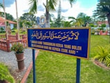 A well-maintained garden with a prominent blue and yellow sign featuring text in both Arabic and Malay. The sign is centered in the foreground, surrounded by green grass, vibrant plants, and various trees. In the background, some decorative elements such as a circular plaque and brick fencing can be seen under a partly cloudy sky.