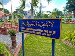 A well-maintained garden with a prominent blue and yellow sign featuring text in both Arabic and Malay. The sign is centered in the foreground, surrounded by green grass, vibrant plants, and various trees. In the background, some decorative elements such as a circular plaque and brick fencing can be seen under a partly cloudy sky.