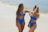 Two women in blue swimwear are standing on a sandy beach near the water. They are smiling and appear to be having a conversation with each other.