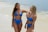 Two women in blue bikinis are standing on a sandy beach near the ocean, smiling and enjoying each other's company. The background shows gentle waves and a clear sky creating a serene beach atmosphere.