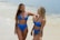 Two women in blue bikinis are standing on a sandy beach near the ocean, smiling and enjoying each other's company. The background shows gentle waves and a clear sky creating a serene beach atmosphere.