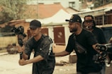 Three men are engaged in filming activities outdoors. One man holds a camera mounted on a gimbal, suggesting a focus on capturing video. Another man stands beside him, appearing to offer guidance or assistance. In the background, another individual holds equipment, indicating a collaborative effort in a filming project. The setting appears to be urban, with buildings and trees visible.