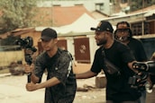 Three men are engaged in filming activities outdoors. One man holds a camera mounted on a gimbal, suggesting a focus on capturing video. Another man stands beside him, appearing to offer guidance or assistance. In the background, another individual holds equipment, indicating a collaborative effort in a filming project. The setting appears to be urban, with buildings and trees visible.