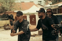 Three men are engaged in filming activities outdoors. One man holds a camera mounted on a gimbal, suggesting a focus on capturing video. Another man stands beside him, appearing to offer guidance or assistance. In the background, another individual holds equipment, indicating a collaborative effort in a filming project. The setting appears to be urban, with buildings and trees visible.