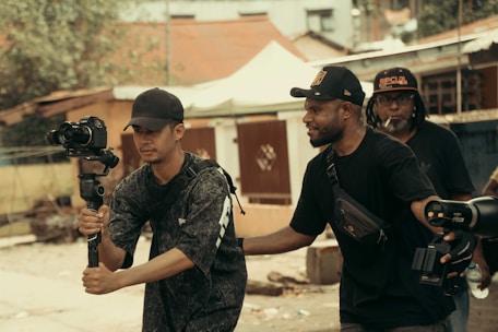 Three men are engaged in filming activities outdoors. One man holds a camera mounted on a gimbal, suggesting a focus on capturing video. Another man stands beside him, appearing to offer guidance or assistance. In the background, another individual holds equipment, indicating a collaborative effort in a filming project. The setting appears to be urban, with buildings and trees visible.