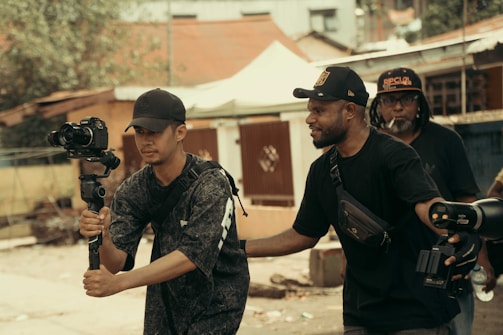 Three men are engaged in filming activities outdoors. One man holds a camera mounted on a gimbal, suggesting a focus on capturing video. Another man stands beside him, appearing to offer guidance or assistance. In the background, another individual holds equipment, indicating a collaborative effort in a filming project. The setting appears to be urban, with buildings and trees visible.
