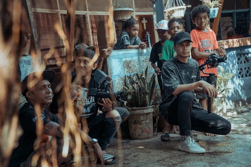 A group of people gathers in an outdoor setting. Some are seated while others stand, and they are surrounded by a rustic backdrop with plants and hanging baskets. One person is holding a camera, suggesting a focus on filming or photography.
