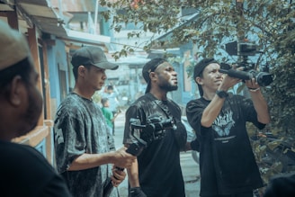 A group of people is gathered in an urban outdoor setting, looking intently at a video camera. Some are holding camera equipment and wearing casual clothing and caps. The background features a street scene with buildings and foliage, creating a busy yet focused atmosphere.