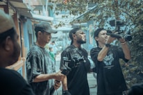 A group of people is gathered in an urban outdoor setting, looking intently at a video camera. Some are holding camera equipment and wearing casual clothing and caps. The background features a street scene with buildings and foliage, creating a busy yet focused atmosphere.