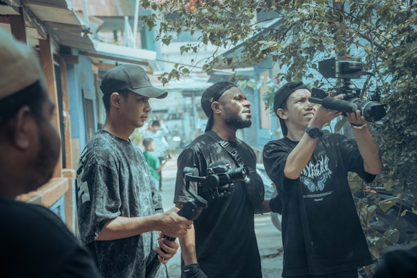 A group of people is gathered in an urban outdoor setting, looking intently at a video camera. Some are holding camera equipment and wearing casual clothing and caps. The background features a street scene with buildings and foliage, creating a busy yet focused atmosphere.