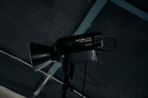 A professional studio light fixture with the label 'NANLITE FS-300B' is mounted and positioned against a dark ceiling with a grid structure. The light has a large conical reflector and appears to be turned off.