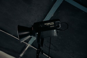 A professional studio light fixture with the label 'NANLITE FS-300B' is mounted and positioned against a dark ceiling with a grid structure. The light has a large conical reflector and appears to be turned off.