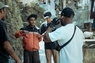 A group of four people are gathered in an outdoor setting beside a wall and some rough terrain. One person in a red jacket appears to be speaking or gesturing, while others listen attentively. They are casually dressed, and the atmosphere seems informal yet engaging.