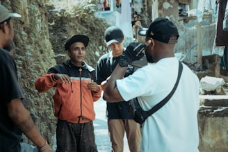 A group of four people are gathered in an outdoor setting beside a wall and some rough terrain. One person in a red jacket appears to be speaking or gesturing, while others listen attentively. They are casually dressed, and the atmosphere seems informal yet engaging.