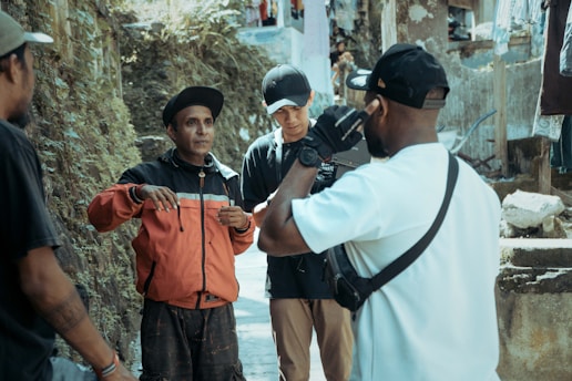 A group of four people are gathered in an outdoor setting beside a wall and some rough terrain. One person in a red jacket appears to be speaking or gesturing, while others listen attentively. They are casually dressed, and the atmosphere seems informal yet engaging.