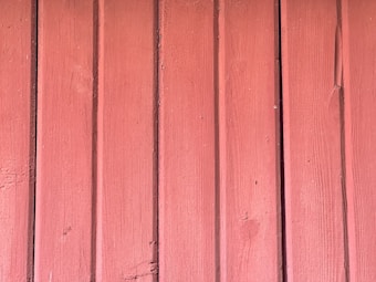 Vertical wooden panels with a worn texture, painted in a muted, rustic red color. The surface shows signs of natural weathering and grain patterns.