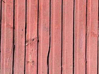 Vertical wooden planks with a reddish hue, displaying natural textures and grain patterns. Some planks have visible knots and imperfections, adding to the rustic appearance.