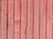 Vertical wooden planks with a reddish hue, displaying natural textures and grain patterns. Some planks have visible knots and imperfections, adding to the rustic appearance.