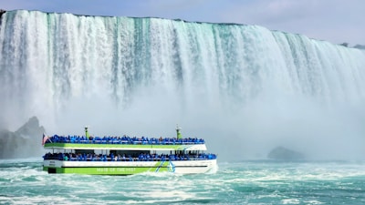 A large waterfall cascades powerfully in the background, with mist rising around it. A boat named 'Maid of the Mist' is filled with people wearing blue ponchos, sailing close to the waterfall. The atmosphere is misty with a sense of adventure.