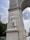 A detailed stone sculpture is inset into a tall, elaborately designed arch or monument. The sculpture features a figure in flowing robes with additional figures in relief behind. The arch is adorned with intricate carvings and emblems. In the background, a cloudy sky and parts of a park with trees and a fountain are visible.