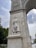 A detailed stone sculpture is inset into a tall, elaborately designed arch or monument. The sculpture features a figure in flowing robes with additional figures in relief behind. The arch is adorned with intricate carvings and emblems. In the background, a cloudy sky and parts of a park with trees and a fountain are visible.