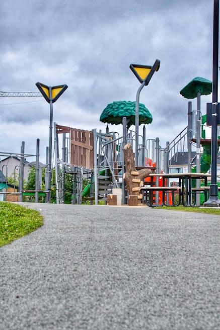 A modern playground featuring various climbing structures, slide, and benches is set under a cloudy sky. The playground includes green, tree-like structures and metal frameworks, with a visible slide and other play equipment. The scene is surrounded by grass and a paved walkway.