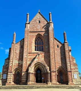 A large, historic red brick and stone church with prominent Gothic architectural features, including tall spires and arched windows. The facade is marked by detailed masonry work, and there are two entrance doors with intricately designed arches.