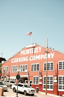 A large red building with a white sign reading 'Monterey Canning Company' stands prominently under a clear blue sky. The building has multiple windows and is bordered by street signs that read 'Cannery Row' and 'Prescott.' An American flag flies atop the structure. Several vehicles, including a white truck, are parked or moving in front of the building. The scene conveys a historic and somewhat nostalgic atmosphere.