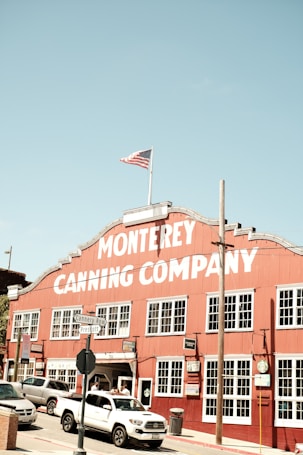 A large red building with a white sign reading 'Monterey Canning Company' stands prominently under a clear blue sky. The building has multiple windows and is bordered by street signs that read 'Cannery Row' and 'Prescott.' An American flag flies atop the structure. Several vehicles, including a white truck, are parked or moving in front of the building. The scene conveys a historic and somewhat nostalgic atmosphere.