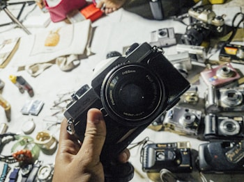 A hand holding a vintage Fujifilm camera with a table in the background displaying various other cameras and photography-related items. The items appear to be laid out for display or sale, suggesting a collection of old cameras and equipment. The setting seems casual and cluttered, with miscellaneous objects scattered around.