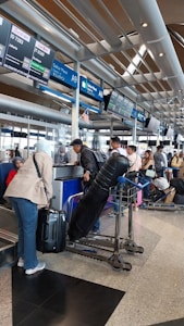 A busy airport check-in area with passengers lining up at the counters. People are handling their luggage, and there are various signs displaying flight information. The environment is crowded, with many travelers preparing for their flights.