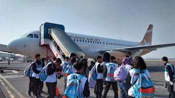 A group of people, many of whom appear to be students with backpacks, are boarding a commercial airplane on an airport tarmac. The airplane is branded with 'SUPER AIR JET' and features a jet bridge or mobile stairs for entry.