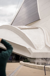 A large, modern architectural structure with sweeping, curved white surfaces and dark glass or solar panels. There is a person in the foreground taking a photo. The architecture appears to incorporate elements of futuristic design.