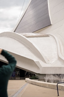 A large, modern architectural structure with sweeping, curved white surfaces and dark glass or solar panels. There is a person in the foreground taking a photo. The architecture appears to incorporate elements of futuristic design.