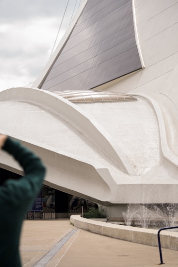 A large, modern architectural structure with sweeping, curved white surfaces and dark glass or solar panels. There is a person in the foreground taking a photo. The architecture appears to incorporate elements of futuristic design.