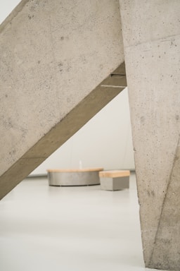 Large concrete structures intersect at an angle, creating a geometric abstract composition. In the background, there are two cylindrical benches, one taller than the other, with a light wooden top, set against a minimalistic, pale floor and wall.